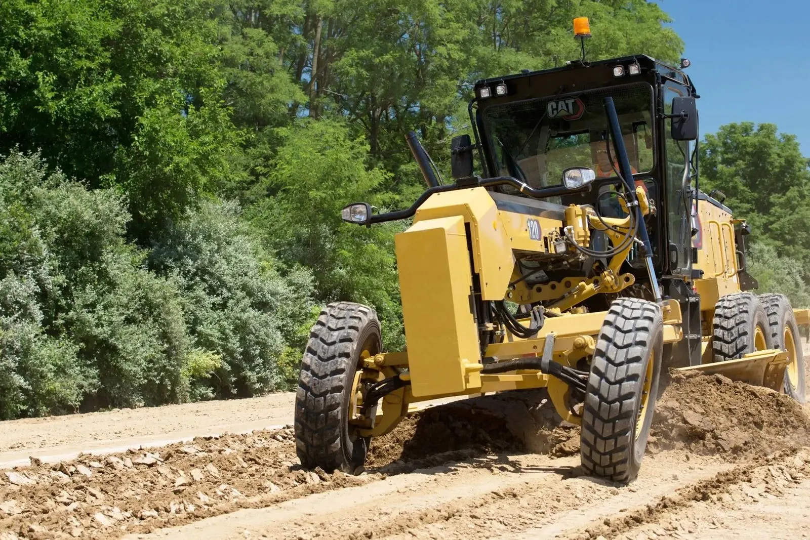 Motor grader leveling a dirt surface with trees behind it